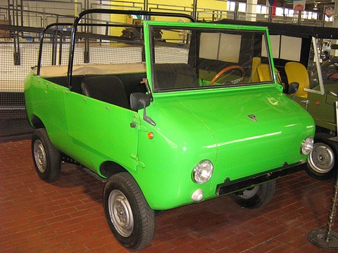 Bright green open four-seat miniature off-roader with round headlamps and a wooden steering wheel, on a brick museum floor. Opens a larger view