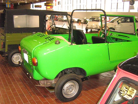 Bright green open miniature off-roader seen from the rear left, with a louvred engine lid and roll bar, on a brick floor. Opens a larger view