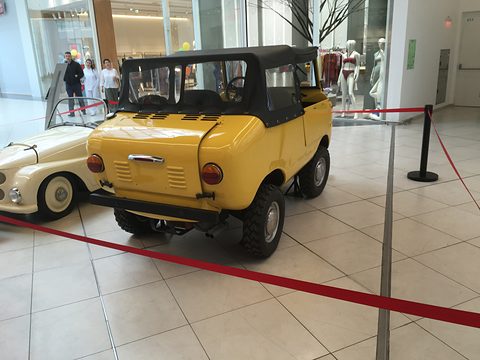 Yellow miniature off-roader with a black soft top seen from the rear right, behind a red rope in a shopping mall. Opens a larger view