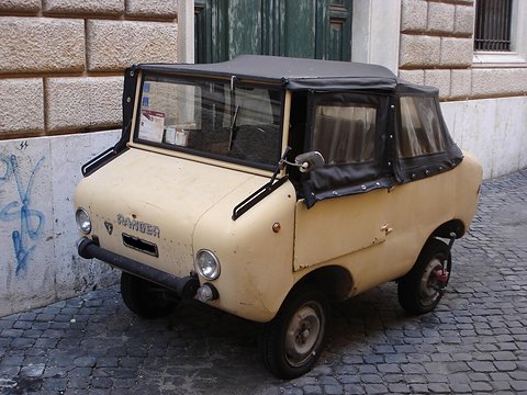 Beige miniature off-roader with a black soft top parked on cobbles beside a stone wall marked with graffiti. Opens a larger view