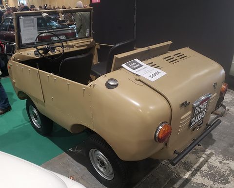 Beige open miniature off-roader seen from the rear right with a dealer plate and price card, on green carpet. Opens a larger view