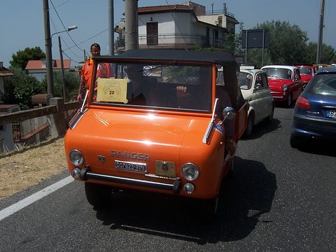 Orange miniature off-roader with a black soft top and a rally number card in its windscreen, leading cars along a road. Opens a larger view