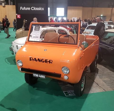 Orange open miniature off-roader with tan seats and RANGER on its nose, Fiat 500s and visitors behind on green carpet. Opens a larger view