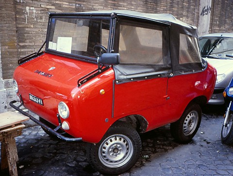 Red miniature off-roader with a tan soft top parked on cobbles against a brick wall, a scooter and car beside it. Opens a larger view