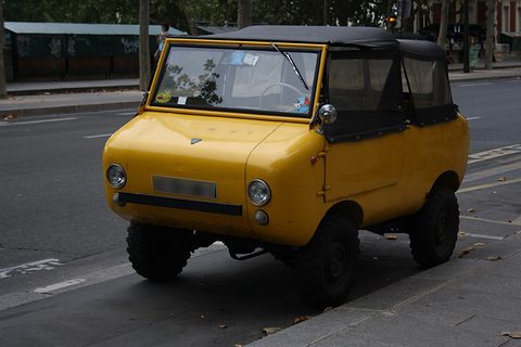 Mustard-yellow miniature off-roader with a black soft top and chunky tyres, parked at a kerb on a wide street. Opens a larger view