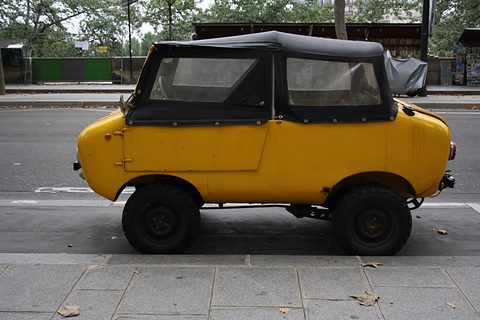 Side view of a mustard-yellow miniature off-roader with a black soft top, parked on a street lined with bookstalls.