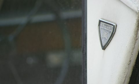 Close view of a chrome shield badge on weathered white paint beside a dark window. Opens a larger view