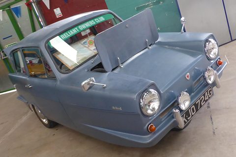 Blue-grey three-wheel saloon with its bonnet raised inside a hall. Opens a larger view