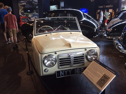 Cream three-wheel convertible facing the camera beside an information board, visitors walking past. Opens a larger view