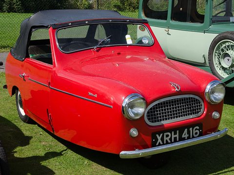 Red three-wheel convertible with a black hood facing the camera, a green vintage car behind. Opens a larger view