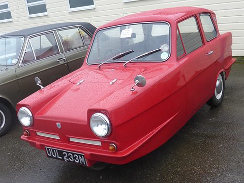 Red three-wheel saloon wet with rain, parked beside a dark saloon car. Opens a larger view
