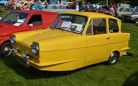 Yellow three-wheel saloon with a display card on the dash, parked on grass by a red van. Opens a larger view