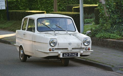 Weathered white three-wheel saloon with round headlamps at a kerb, hedges behind. Opens a larger view