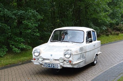 White three-wheel saloon with peeling paint driving on a brick-paved lane through woods. Opens a larger view