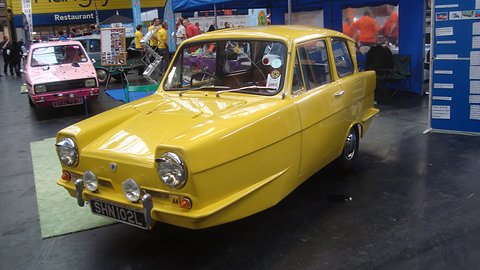 Yellow three-wheel saloon with spot lamps on a dark hall floor, a pink car behind. Opens a larger view