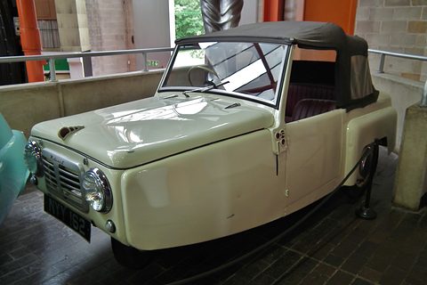 Cream three-wheel convertible with a grey hood and slatted grille behind a museum rail.
