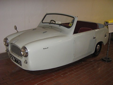 White open three-wheeler with a mesh grille and wrap-round windscreen on a brick museum floor.
