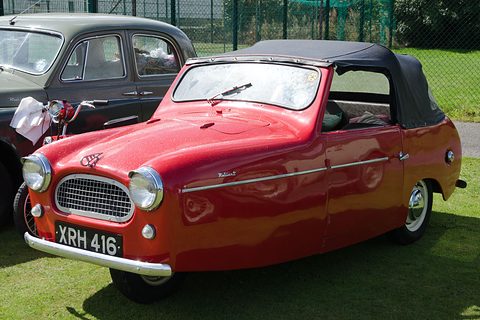 Red three-wheel convertible with a black hood and mesh grille on grass by a fence.