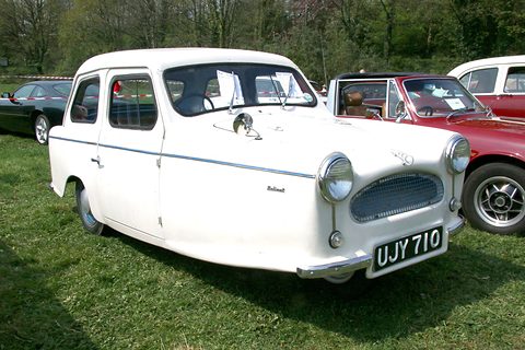 White three-wheel saloon with a blue side stripe parked on grass among classic cars. Opens a larger view