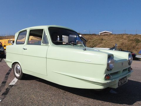 Pale green three-wheel saloon with round headlamps on a tarmac car park under blue sky. Opens a larger view