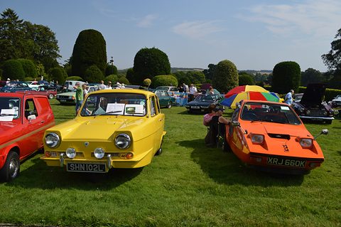 Yellow three-wheel saloon and a wedge-shaped orange three-wheeler on a lawn with clipped yews. Opens a larger view