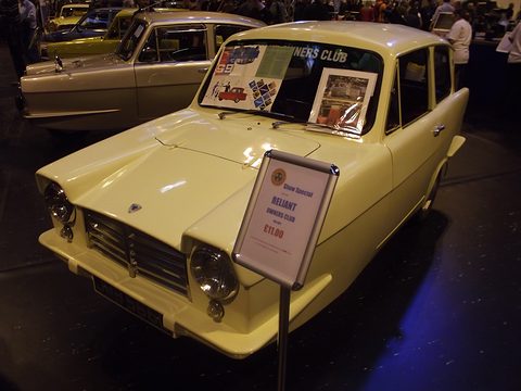 Cream three-wheel saloon with a for-sale board in front, in a dim exhibition hall. Opens a larger view