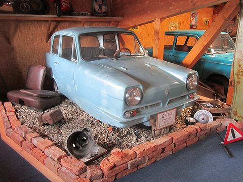 Pale blue three-wheel saloon in a mock garage with gravel, bricks and a warning triangle. Opens a larger view