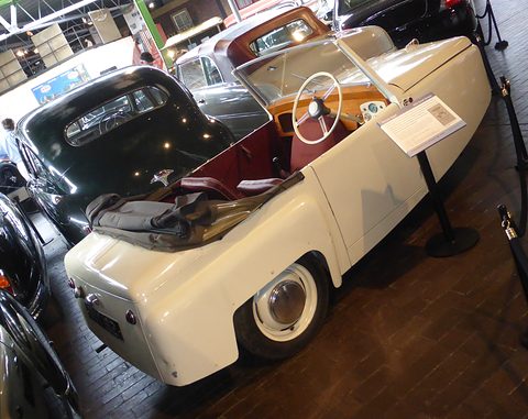 Cream open three-wheeler with red seats, seen from the rear quarter among dark museum cars. Opens a larger view