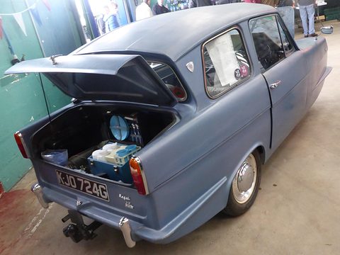 Blue-grey three-wheeler seen from the rear quarter with its boot lid raised over tools. Opens a larger view
