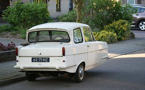 White three-wheel saloon with a reverse-rake rear window parked at a kerb by shrubs. Opens a larger view