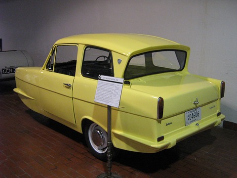 Yellow three-wheel saloon with a reverse-rake rear window and an information stand beside it. Opens a larger view