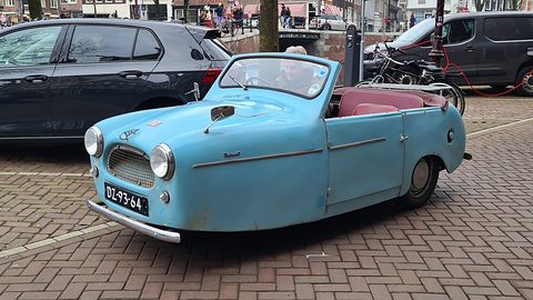 Weathered pale blue open three-wheeler on brick paving beside a dark hatchback.