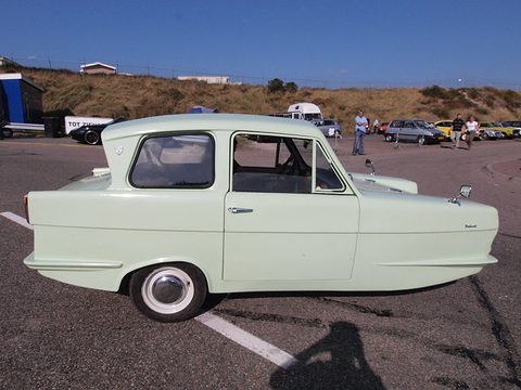 Pale green three-wheel saloon in side view on a car park, dunes behind. Opens a larger view