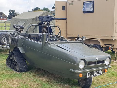 Grey open three-wheeler on rubber tracks with a mock machine gun, beside a military trailer. Opens a larger view