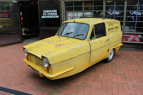 Weathered yellow three-wheeled van with a slatted grille nose on brick paving outside a showroom. Opens a larger view