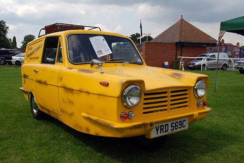Yellow three-wheeled van with a slatted nose and roof rack on grass, a brick pavilion behind.