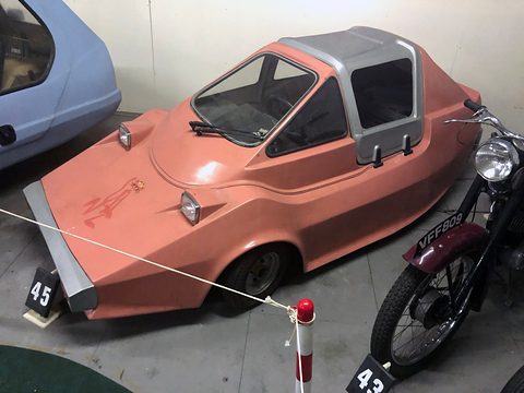 Pink wedge-shaped microcar with a grey roof, behind a rope beside a vintage motorcycle.