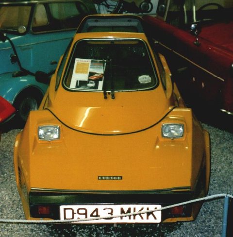 Orange wedge-shaped microcar head-on in a museum line-up, a red car beside it. Opens a larger view