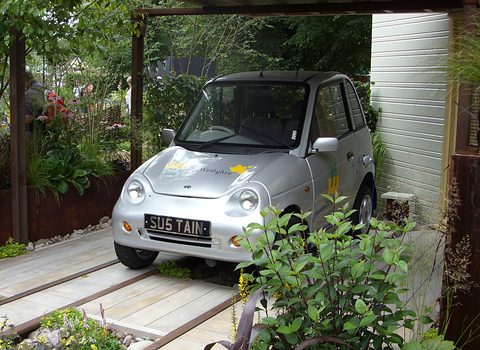 Silver microcar with a SUSTAIN number plate parked on a timber deck among plants under a wooden pergola. Opens a larger view