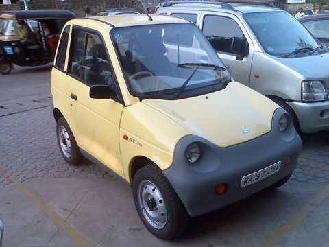 Pale-yellow microcar with grey lower panels and round lamps parked beside a silver hatchback, an auto-rickshaw behind.