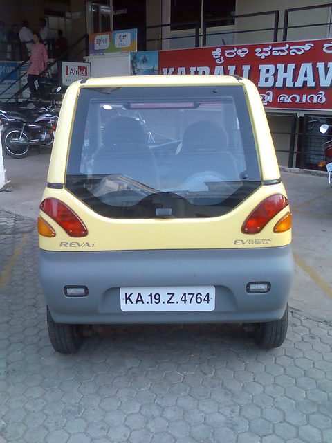 Rear of a pale-yellow microcar with grey bumper, round tail lamps and a white plate, motorbikes and shop signs behind. Opens a larger view