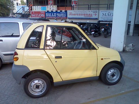Side view of a pale-yellow microcar with grey lower panels and steel wheels in a car park, shop signs behind. Opens a larger view
