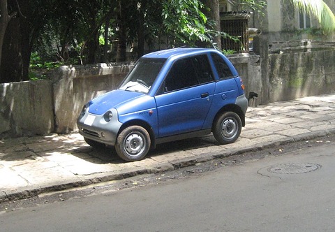 Small blue tall two-door car on a pavement under palms and trees beside a low garden wall. Opens a larger view