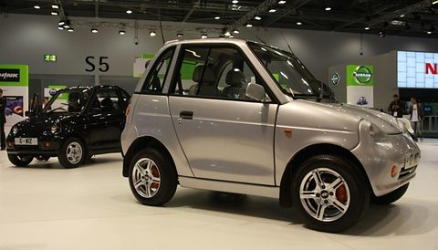 Silver tall two-door microcar with alloy wheels on a white stand under show lights, a black example behind. Opens a larger view
