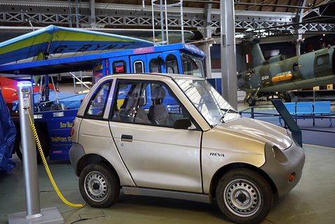 Champagne-coloured microcar connected by a yellow cable to a charging post in a hall with vehicles behind. Opens a larger view