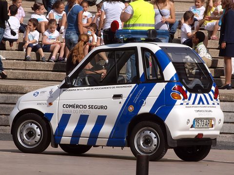 White microcar with blue chequered police stripes and a roof light, spectators seated on steps behind. Opens a larger view