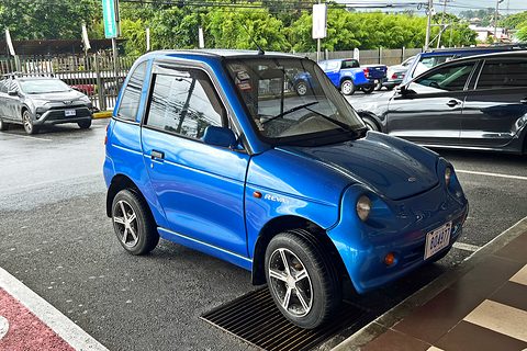 Metallic blue tall two-door microcar with alloy wheels parked over a drain grating on a wet car park. Opens a larger view