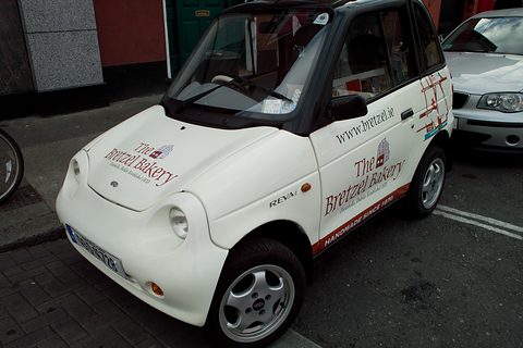 White microcar lettered ‘The Bretzel Bakery’ on bonnet and doors, parked at a kerb on a city street. Opens a larger view
