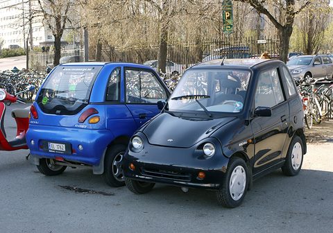 Two short tall microcars, one blue seen from behind and one black from the front, by rows of bicycles. Opens a larger view