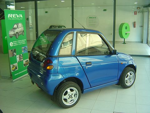 Blue microcar with alloy wheels on a tiled showroom floor beside a green REVA display stand. Opens a larger view